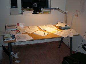 A desk covered in notes, articles, books, used for research. The desk also has a lamp.