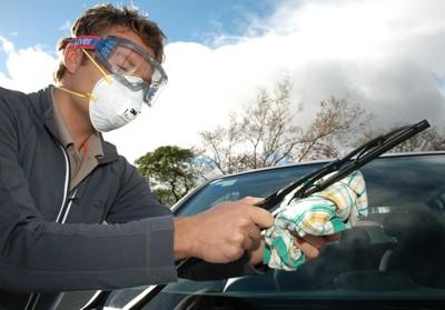 A man wearing goggles and a mask carefully cleans volcanic ash off a windshield.