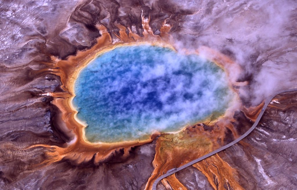 An aerial view of Grand Prismatic Spring, which is steaming and bright blue and ringed by rainbow colors of reds, oranges, and yellows caused.