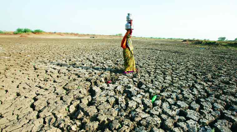 A woman is shown carrying water on her head while crossing dried, deeply cracked ground.