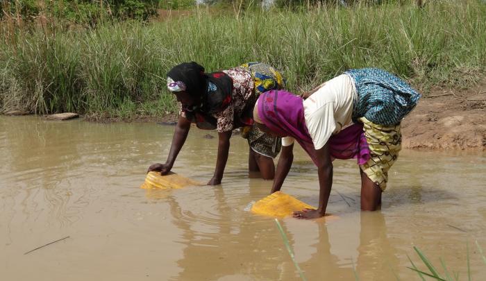 This photo shows two women in Ghana scooping brown water as they are standing in it.