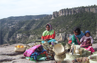Two Tarahumara, one holding a baby, sit weaving baskets near a cliff edge in Mexico