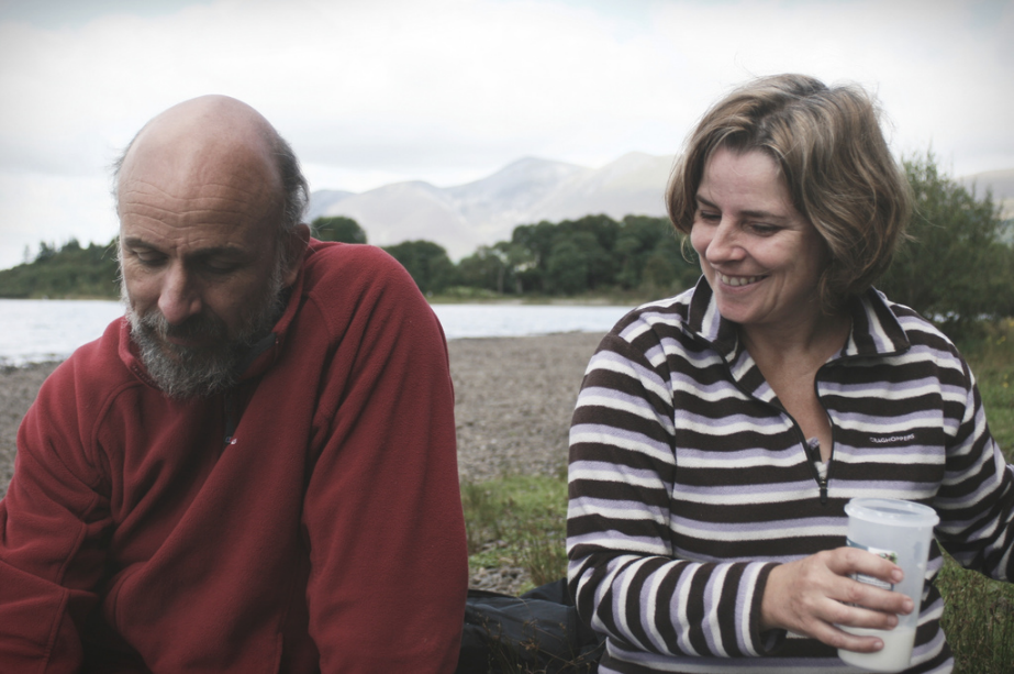 two people outside, one holding a drink with lake and mountains in the background