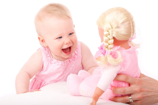 A toddler in pink smiling at a doll with blonde hair dressed in pink.