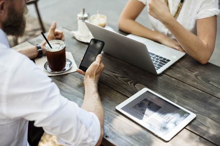 person with laptop, tablet on table, and someone sitting across the table with iced coffee and holding phone