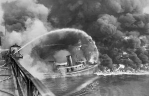 Photo of a river and a boat on fire, with firefighters spraying water on the fire