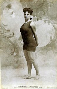 A black and white vintage photo shows a woman is standing in one piece bathing suit. She is standing with her hands behind her back.