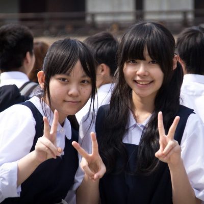 Two young Asian girls in school uniforms are making the peace sign with their fingers.