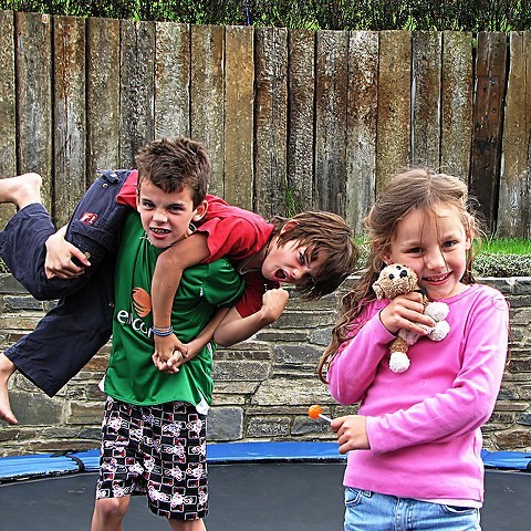 Two boys are playing and one boy is carrying the other boy over his shoulders. A girl is in the foreground smiling and holding a stuffed animal. 