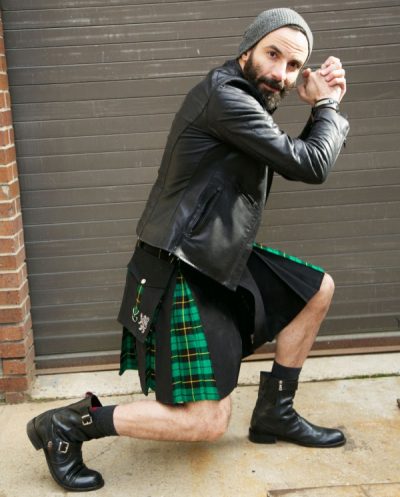 A man is bending at the knees with his hands gripped in a victory pose. He is wearing a black leather jacket and a green and black kilt.