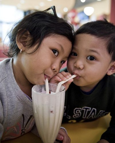 Two Asian children are sharing a milkshake; there is one ice cream shake and the children are using two separate straws.