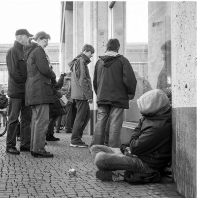 A homeless person sits against a wall, their face is covered by a hoodie; onlookers around the person are ignorning them.