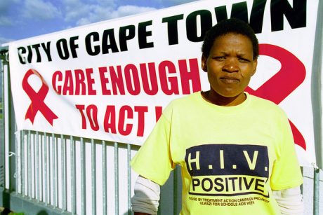 A Black woman in a yellow shirt that reads HIV positive, stands in front of a sign that reads City of Cape Town: Care Enough to Act.