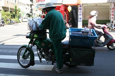 A courier is getting on his motorcycle. The materials being delivered are in a large container at the back of the motorcycle..