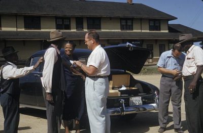 A group of four Black men and women are receiving items from two males. There is a car behind the group of people and the truck is open showing boxes.