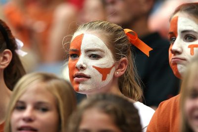 A college student in a crowd. Her face is painted orange and white. The University of Texas Longhorn symbol is painted in orange on her cheek that is painted white.