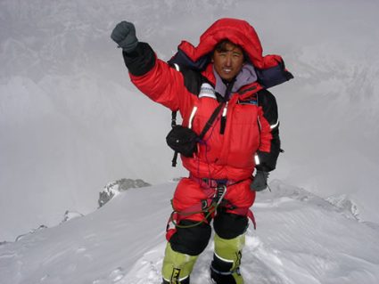 A sherpa man in a bright red jacket is standing on a snow covered mountain outcrop. His hand is raised in the air in a sign of triumph.