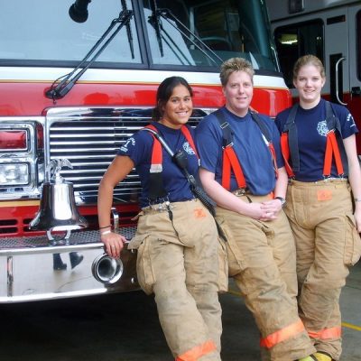 Three female firefighters are standing in front of their fire truck.