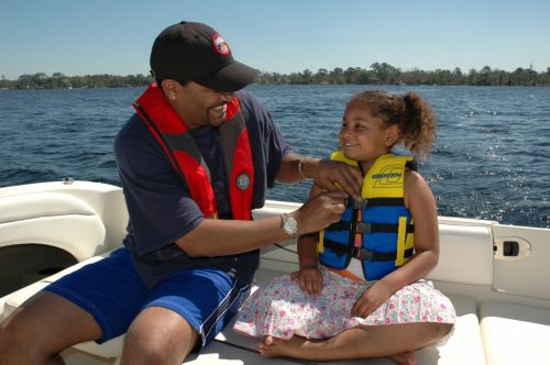 A man and child are on a boat and the man is helping the female child with her lifevest.