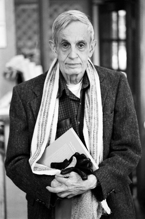 Black and white photo of John Nash, Nobel Prize Winner. He is standing with his arms crossed at his waist and he is holding a book between this arms and his body.