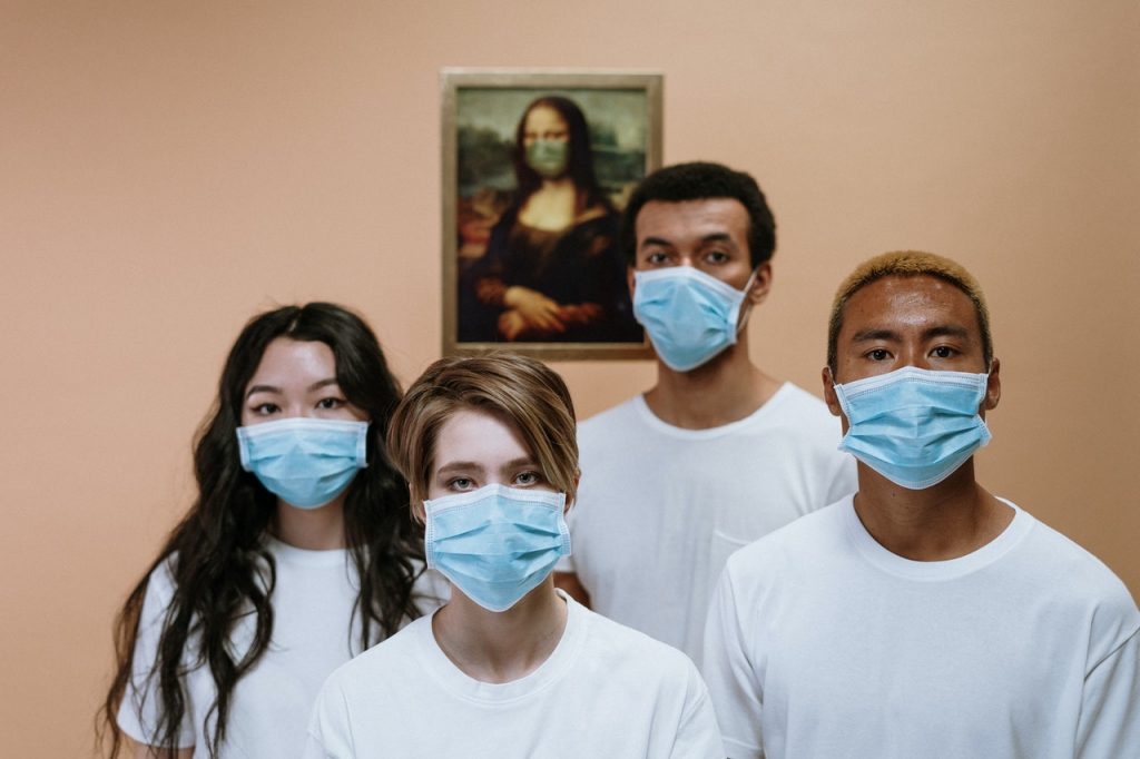 Four people standing in a room wearing hospital masks and white T-shirts. A picture of the Mona Lisa is on the wall behind them wearing a mask.