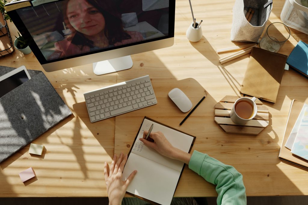 hands writing in notebook at a desk. Desktop computer on desk shows a virtual meeting