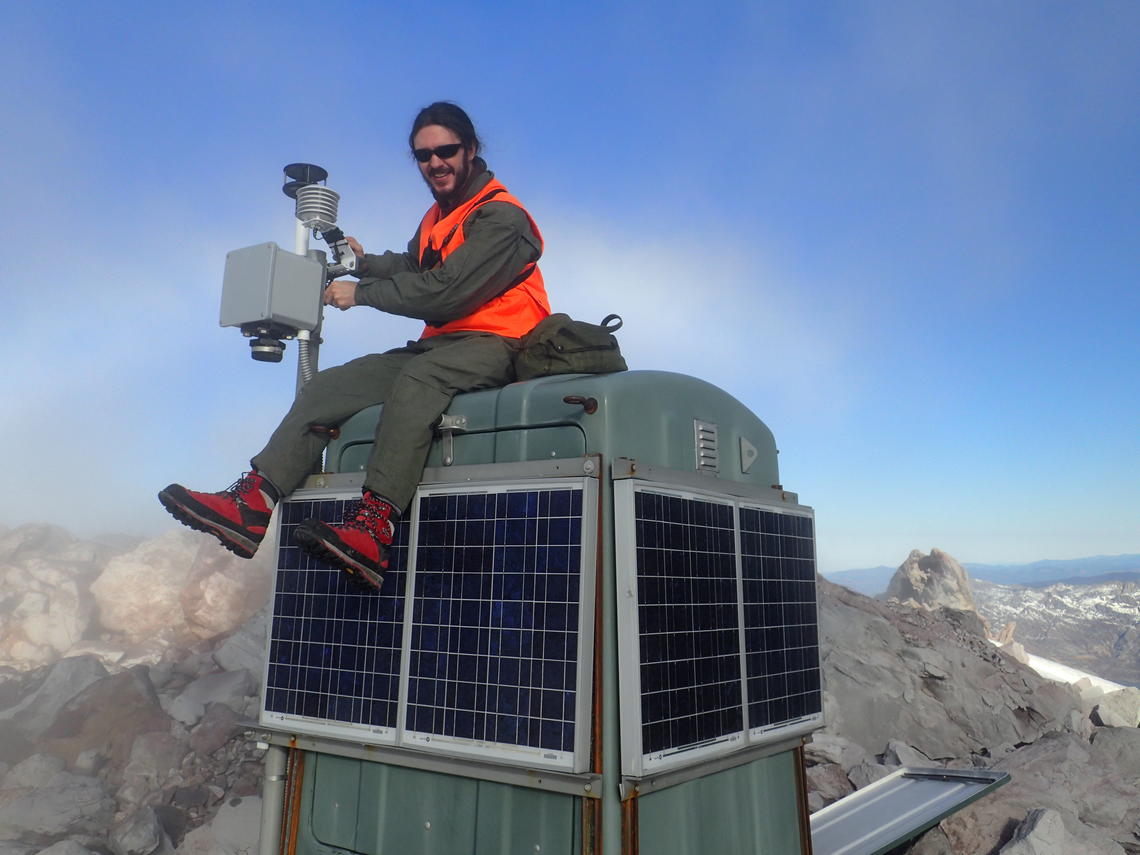 Large gas monitoring station with solar panels at Mount St. Helens with a geologists sitting on top of it for scale.