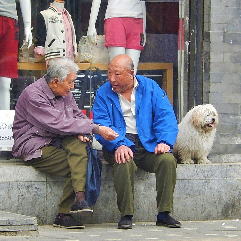 Two older Asian men sitting on a raised sidewalk talking.