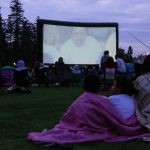 families sit outdoors to watch a movie on a large screen