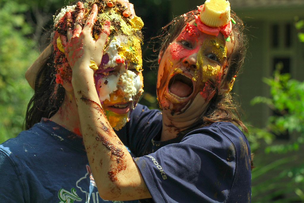 a women, mouth open, rubs cake on her friends head