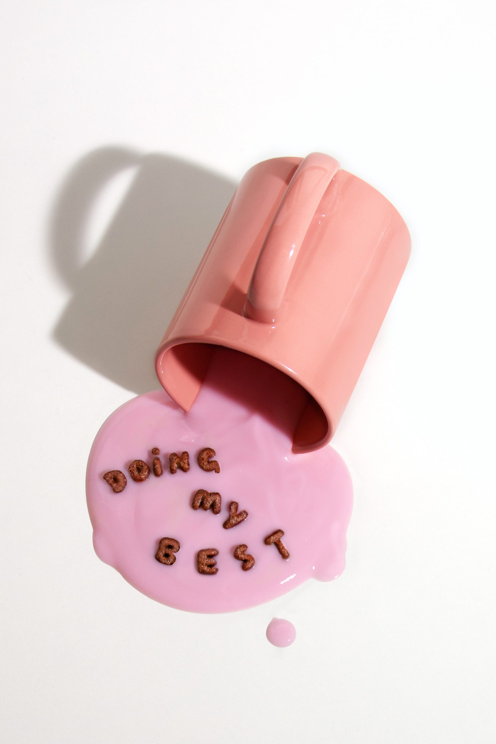 A pink cup spilling the pink liquid on the white surface and some alphabetical red cereal that are arranged to build a sentence, “Doing my BEST.”