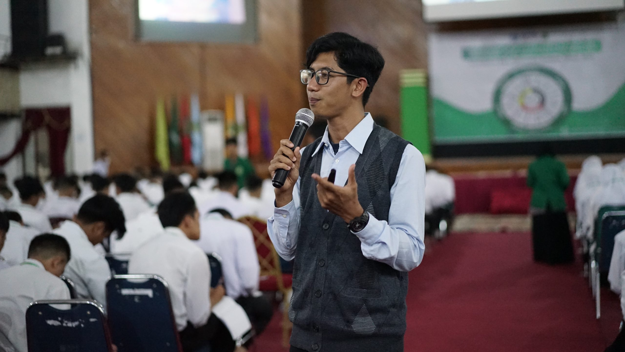 A man speaking with a microphone on his right hand and a pen or slide clicker on his left hand. He is standing and looking at the audience in a room full of students.