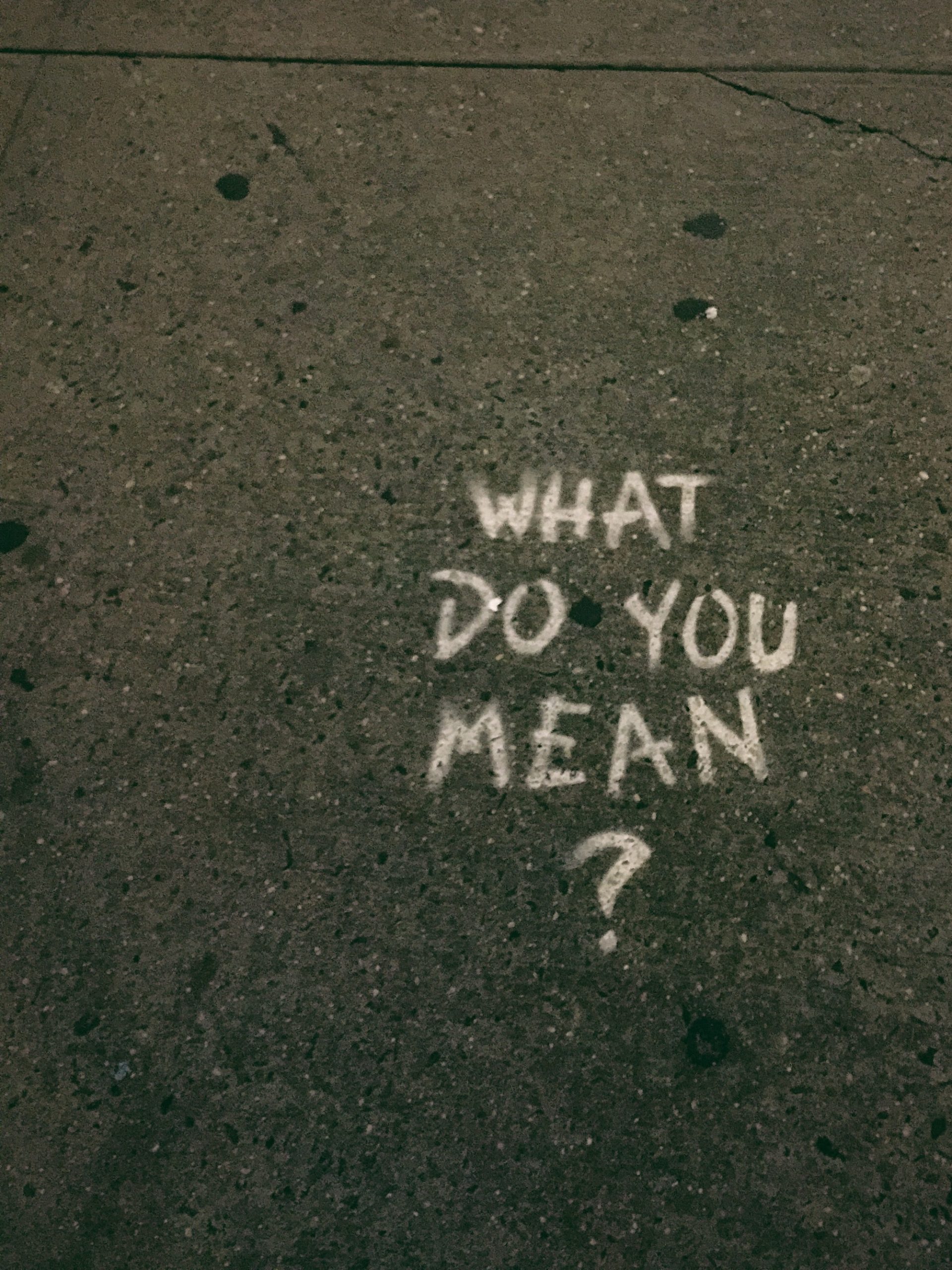 A white chalk written on the street that says, “What do you mean?”.