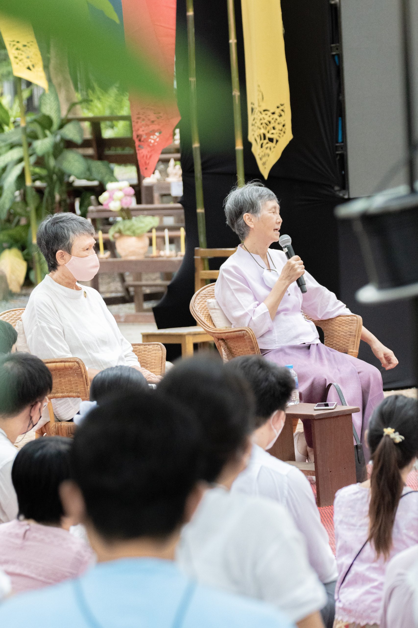 Two women sitting on the chairs, one woman speaking holding a microphone in an outdoor environment, and the audience are sitting on the floor on the carpet.