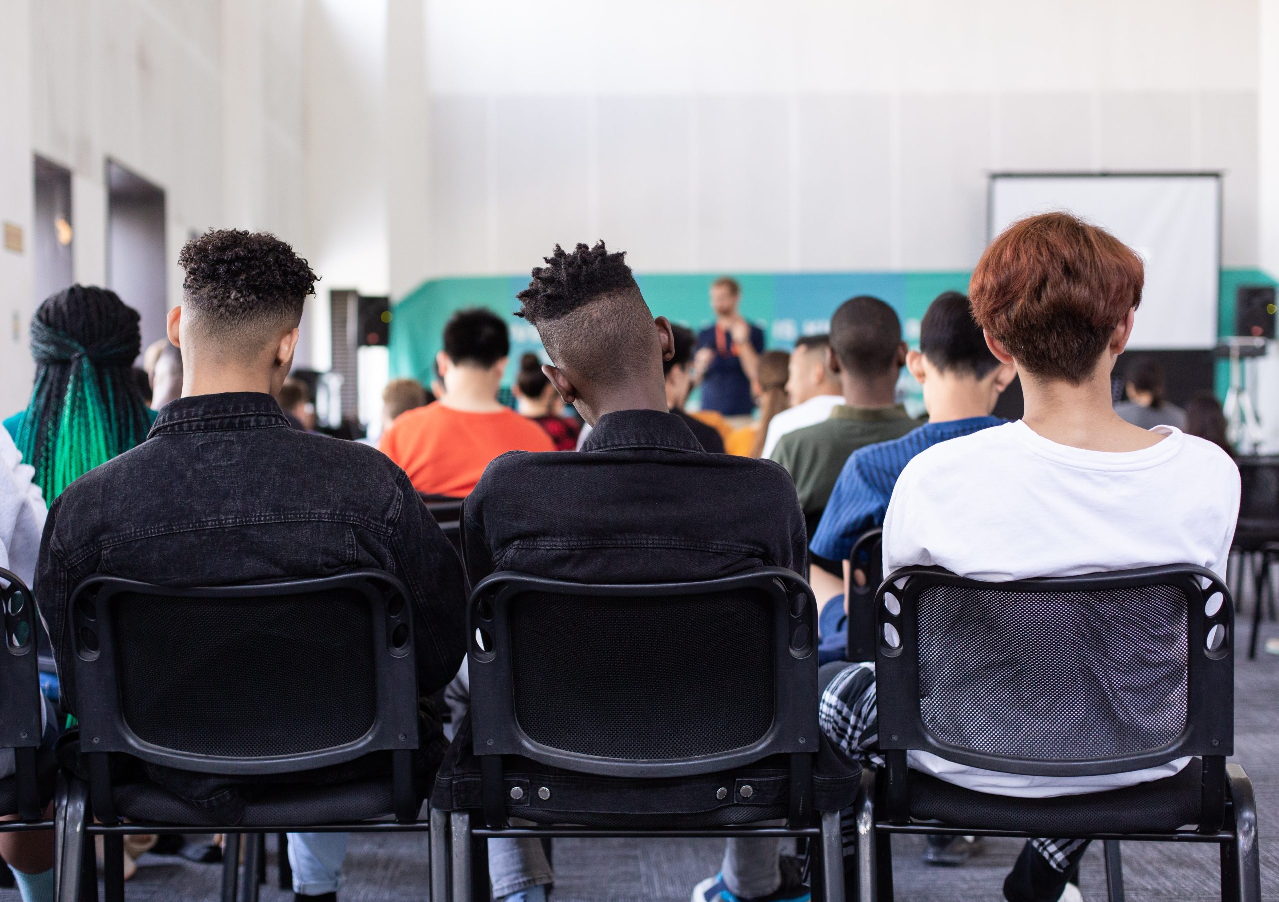 Some students sitting on the chair in the classroom and the teacher standing in front of the class facing students.