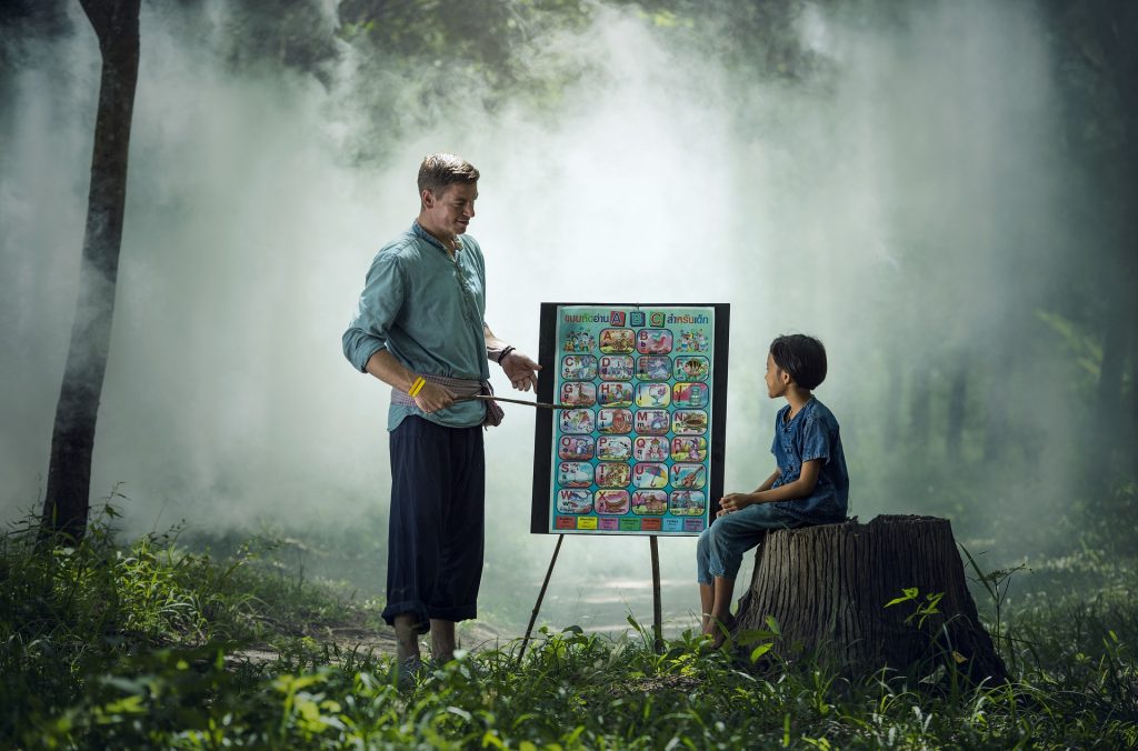 A teacher standing in a forest using a stick to point at a diagram of the alphabet while a child is watching and sitting on a tree stump.