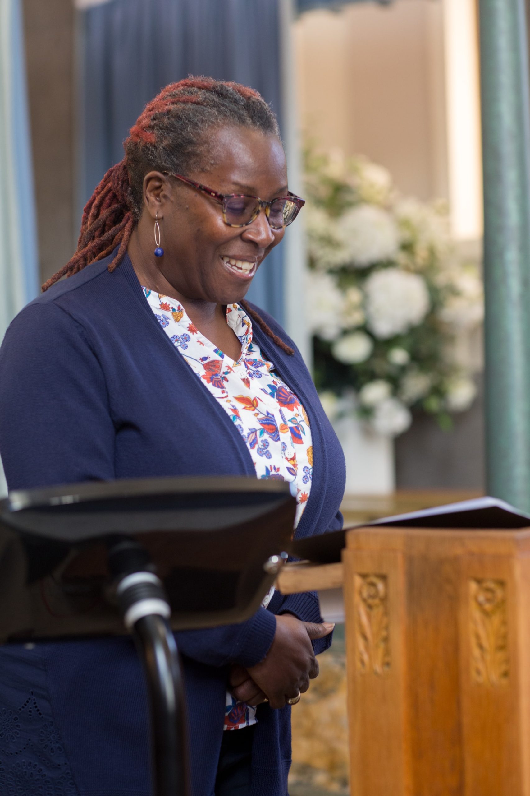 A woman wearing a colorful blouse stands at a lectern at a funeral ceremony.
