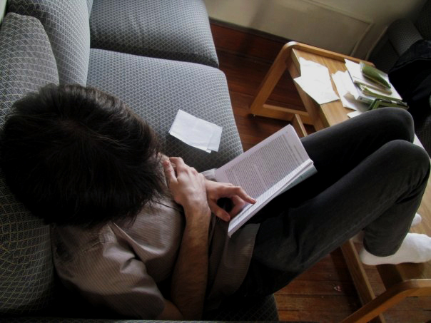 A student sits on a couch reading a textbook. Papers and notes are scattered on the coffee table and couch cushions.