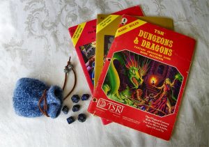 A fuzzy blue drawstring pouch lays on a table with dark blue polyhedral dice spilling out. A the 1981 Dugeons & Dragons rules booklet lays fanned out on top of two adventure module expansion booklets.