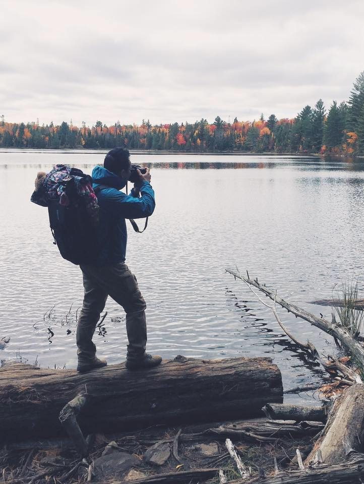 Taekyom Kim stands on a log beside a lake with a large backpack, taking a picture