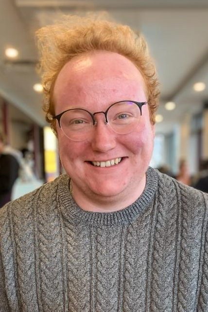 Caleb, a white man with curly red hair and large round glasses smiling.