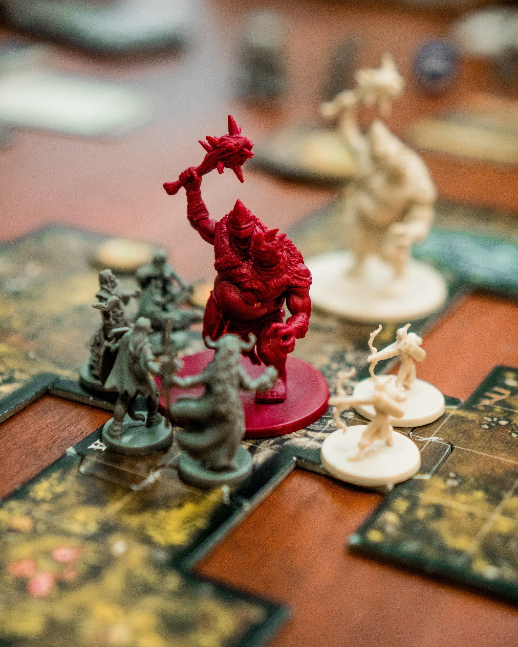 A large red monster figurine has a weapon raised above his head. The monster is surrounded by 7 small figurines with bows, spears, and swords. The figurines are placed on map tiles which describe and map out the environment.