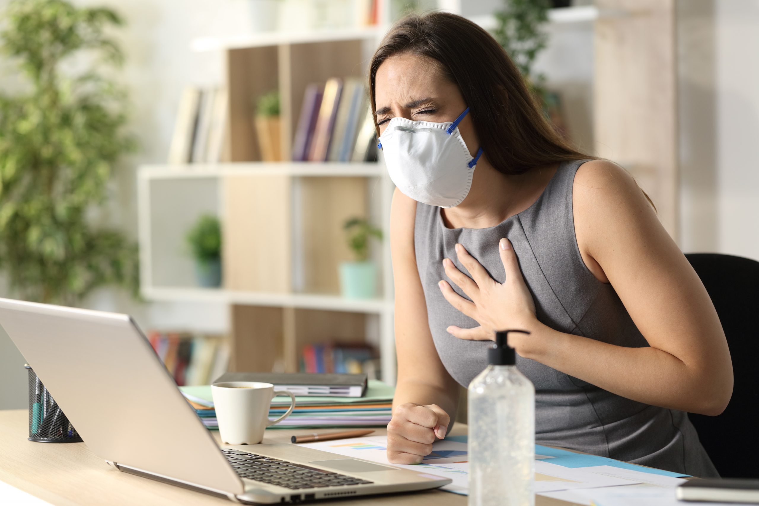 Image of a masked woman sitting at office desk, with hand to chest in possible duress