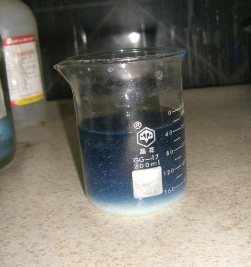 A glass beaker with a blue solution contained within, precipitate has formed in the solution