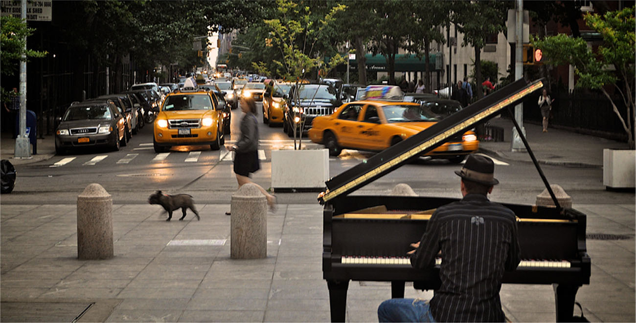 A photograph shows a person playing a piano on the sidewalk near a busy intersection in a city